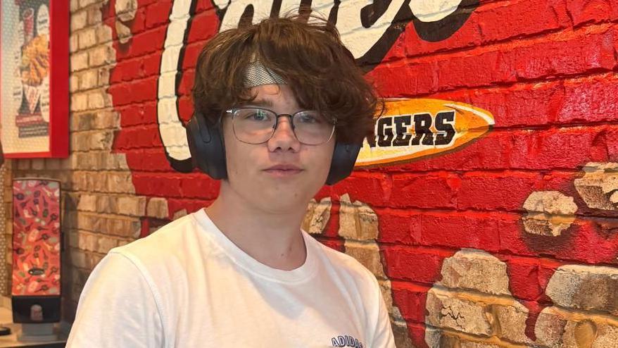 Sam Bailey's son Tommy looking at the camera in front of a red wall with lettering in a fast-food restaurant. He's wearing glasses and headphones.