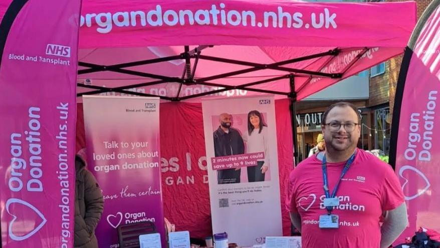 Ben Cronin stands in front of pink stall that offers information about organ donation during an event. He wears a pink top to match the stall. There are boards with information about organ donation behind him as well as a pink gazebo.