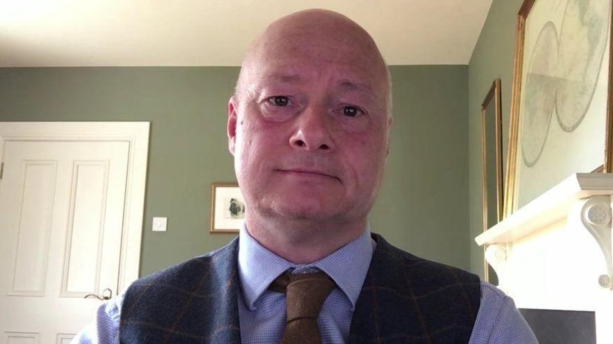 Paul Bartholomew of Ulster University. He is a bald man and is wearing a blue  shirt, brown tie and tweed waistcoat. He is sitting in a room with green walls and a white door