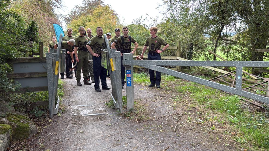 A group of about ten men on a countryside track standing and facing a study metal gate