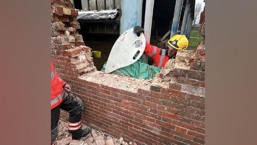 Fire officer shielding a sedated and covered horse. the brick wall has been partly removed.  