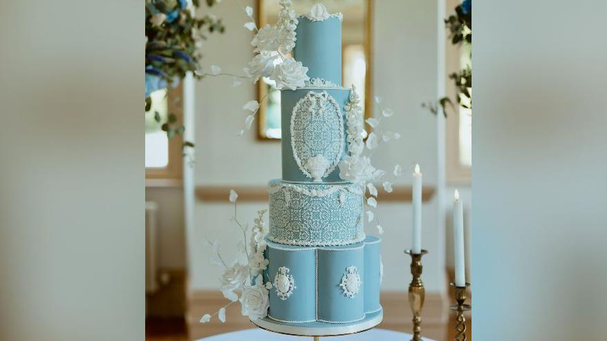 The picture shows a pale blue cake with a lace design. The cake has four tiers.  It is placed on a table and is surrounded by candles and flowers.