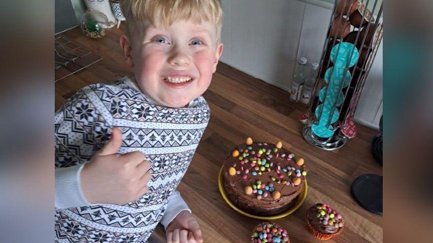 Alfie in a black and white top smiling and doing a thumbs up sign. He is smiling broadly lying on a brown floor beside chocolate cake decorated with colourful chocolate buttons. 