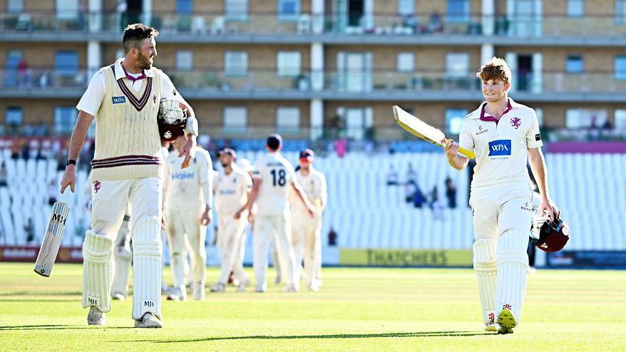 Craig Overton (left) holds his bat in his right hand and helmet under his left and looks at Thomas Rew (right), as they walk off the pitch.