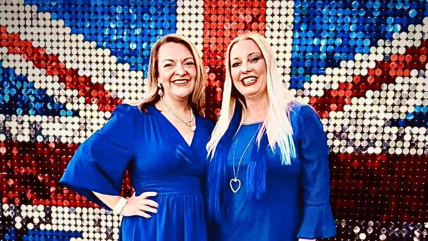 Two women wearing blue dresses standing in front of a union jack backdrop
