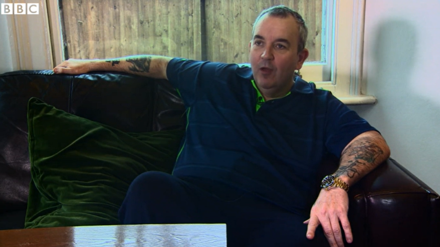 A man sitting on a sofa, with patio doors behind him. He has short greying hair and is wearing a blue polo shirt.