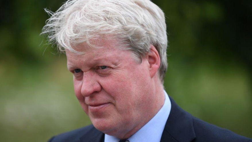 Earl Spencer with greying hair and wearing a dark blue blazer with pale blue  shirt and tie. He is outdoors and and there is greenery behind him.