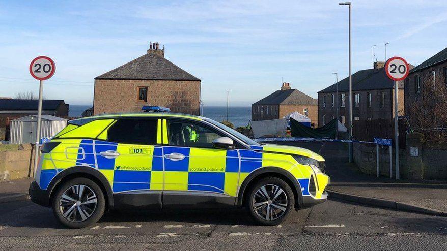 Police car with yellow and blue markings at scene of incident, with police tape in background, and two 20mph speed limit signs, and blue sky.