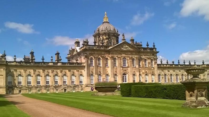 A huge stone-coloured stately home with green lined grass and square hedges on a blue sunny day.