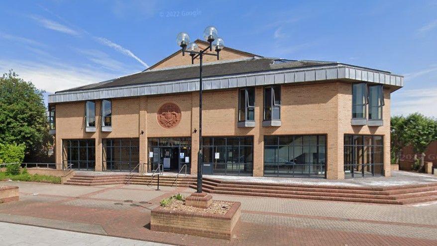 Lincoln Magistrates' Court. The building is made from pale-coloured bricks. It has large windows with brick paving outside and raised flower beds.