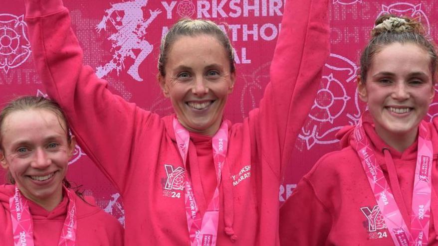 The first, second and third-placed runners in the women's race wear pink hoodies and their race medals on pink lanyards. They are smiling and the winner is raising her arms. They stand in front of a pink Yorkshire Marathon banner.