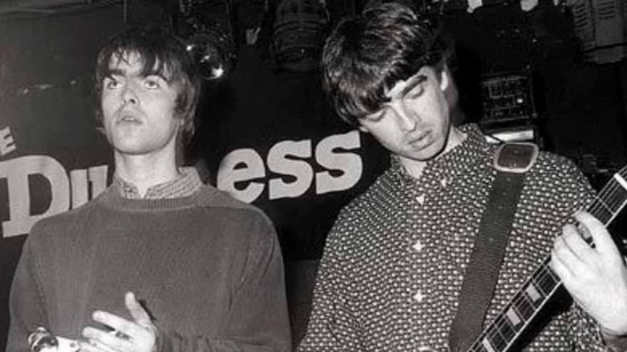 Two men on a stage, Liam and Noel Gallagher. The man on the left, Liam, has a shaggy haircut and is wearing a shirt underneath a jumper. The man on the right, Noel, is playing a guitar and is wearing a spotted shirt. In the back there is a black sign with white writing saying Duchess.