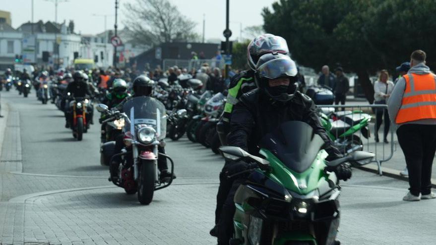 Motorcyclists travel down a road in single file during the Shakedown event. Crowds watch on from the side of the road with other motorbikes parked up in front of them. An event marshall in an orange hi-vis jacket watches the crowd.