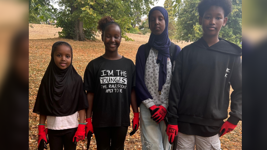 Four children stand in a row in a park. There are three girls and one boy. Two of the girls are wearing headscarves. They all wear read gloves and hold litter pickers. 