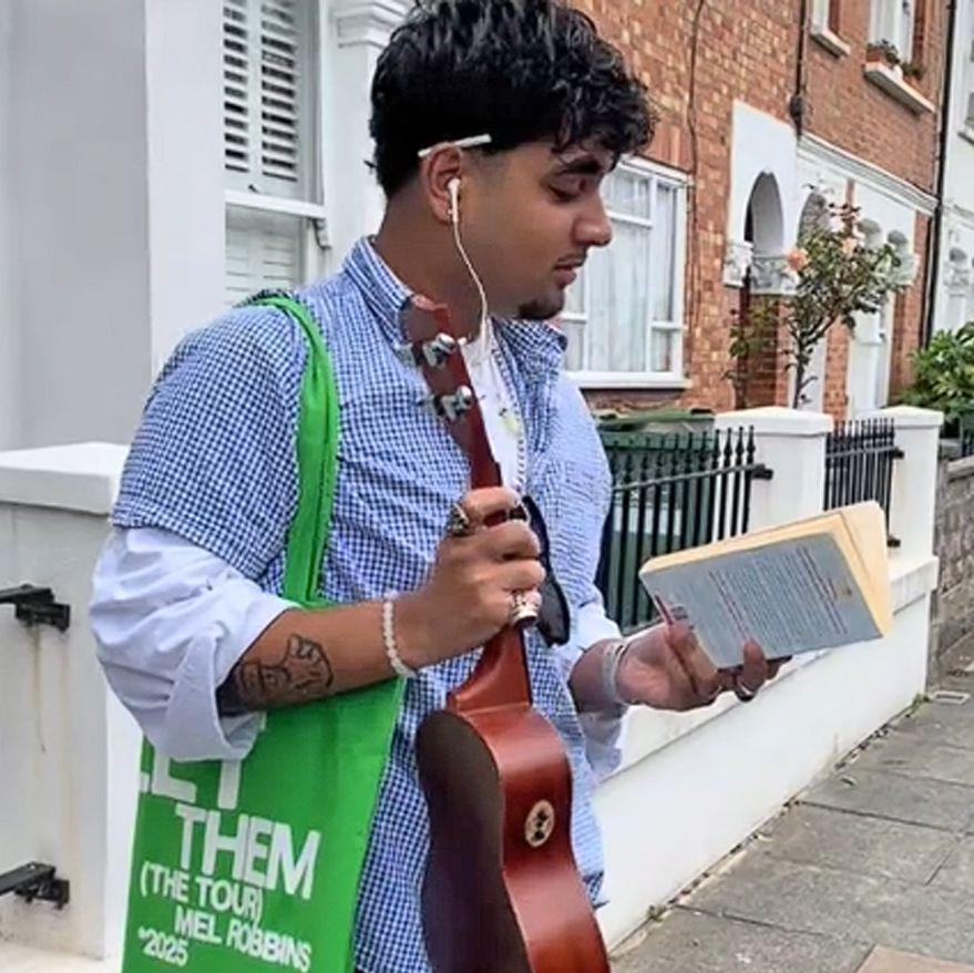 A young man poses on a street as he reads a feminist book in one hand and clutches a ukulele in the other. He has a tote bag on his shoulder as well as a Labubu and vintage cap hanging from his belt loops.