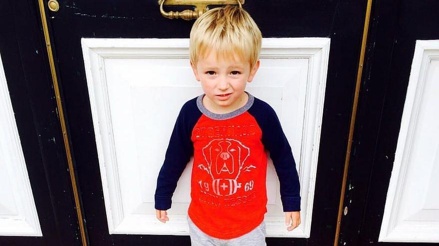 Tommy in a red top as a young boy, standing in front of a door.