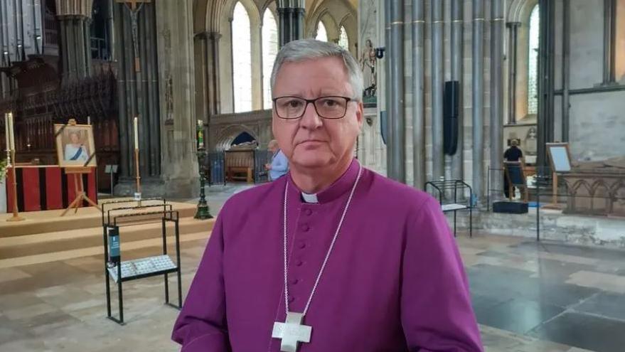 The Bishop of Salisbury stands in Salisbury Cathedral in purple clergy garments with a large cross round his neck.