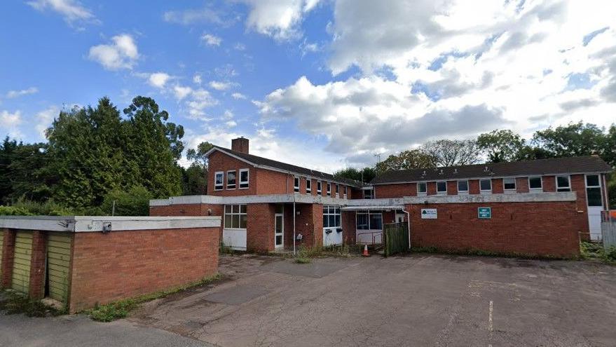 A Google streetview image of the existing building. It is a large, two-storey red-brick building, and appears significantly run down. There are lots of weeds growing in a large parking area, and at least one of the garage doors in warped.
