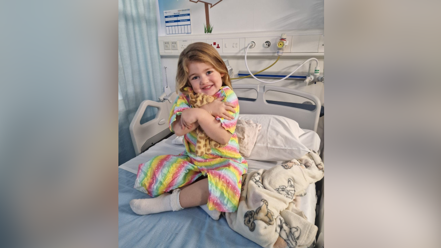 Poppy before her operation in a hospital bed cuddling a soft toy and smiling