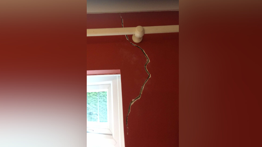 A large, winding crack is seen snaking down a living room wall, beginning at the ceiling and ending around halfway down the wall near a window. A wooden curtail rail runs along the top. The crack is quite wide, with a visible gap between the two sides. 