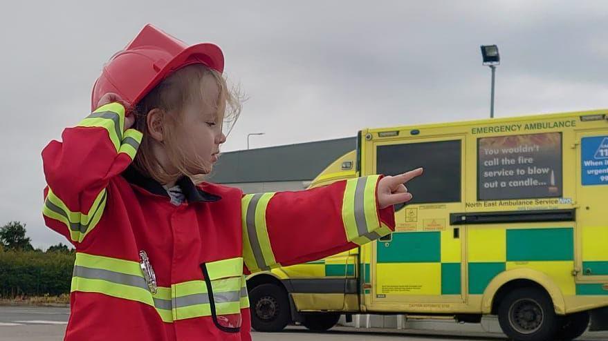 Beatrix is wearing a red and yellow fire fighter uniform, including a hard hat and a badge. She is pointing at something in the distance. An ambulance can be seen parked in the background.