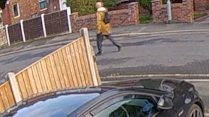 A still of a man in an orange coat wearing a black rucksack walking along the middle of the road on a residential street. 
