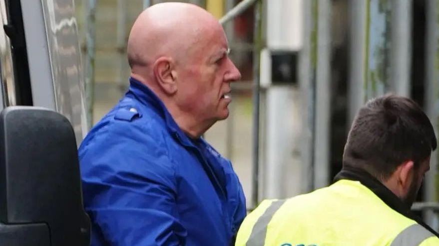 Dyn yn cerdded allan o ddrws fan tuag at ddrws llys. Ochr ei ben sydd i'w gweld, gyda'i glust dde yn wynebu'r camera. Mae'n gwisgo siaced glas.