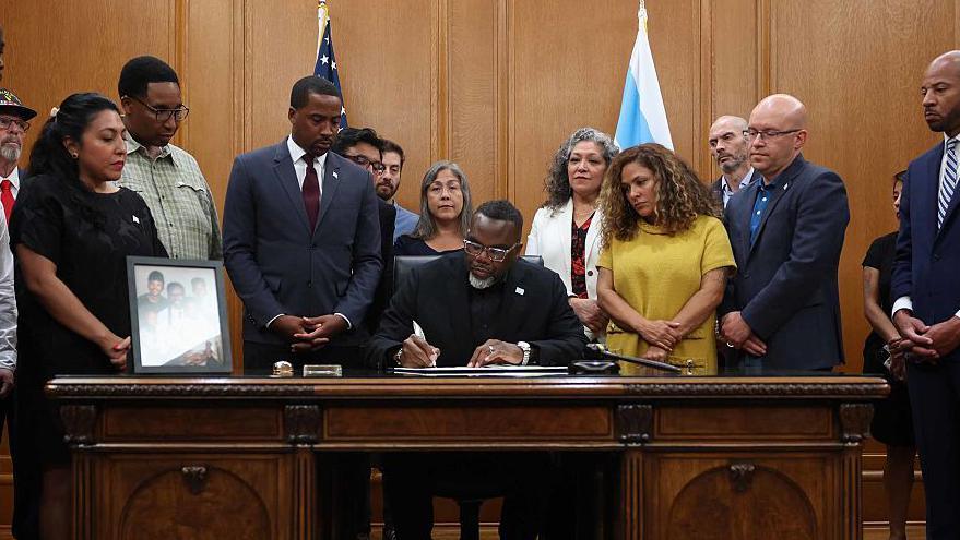 Mayor Brandon Johnson sitting behind a brown desk signs an executive order. He is flanked on his left, right and behind him by officials.