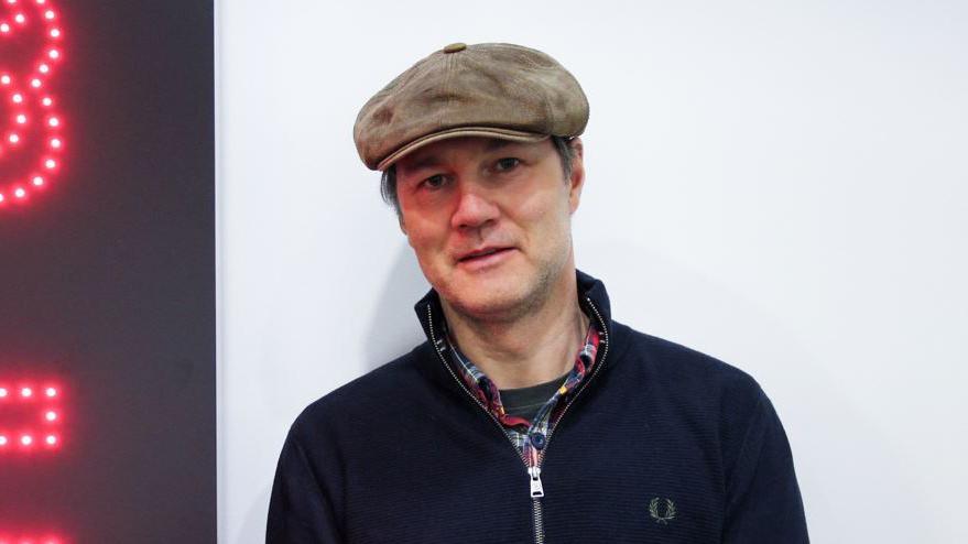 David Morrissey smiles at the camera in BBC Studios, wearing a brown flat cap and Fred Perry navy jacket