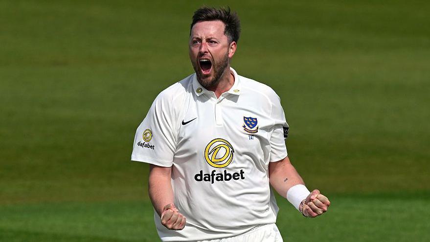 Ollie Robinson, wearing a white Sussex shirt, roars and clenches both of his fists in front of him to celebrate a wicket
