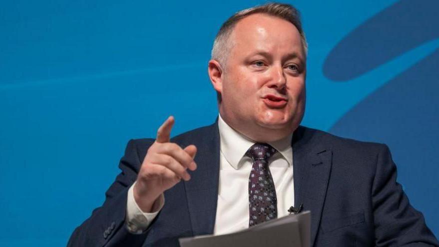 Darren Millar is delivering a speech to an audience. His right finger is pointing upwards. He is wearing a suit, tie and shirt, and is stood before a dark background.