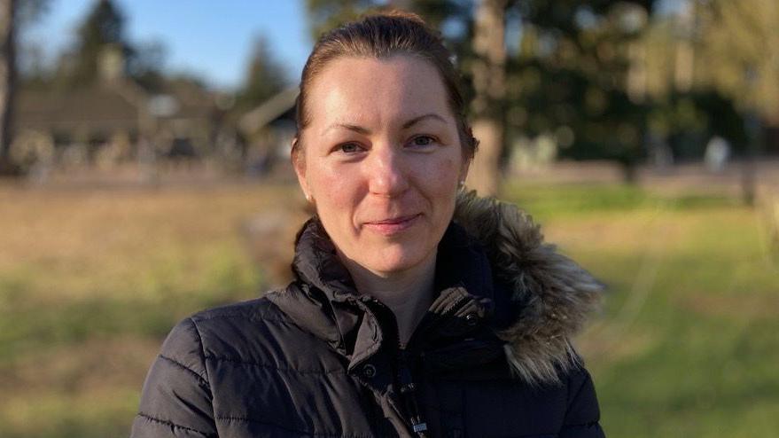 Woman wears a black puffer coat with black fur trim. She is standing in a park.