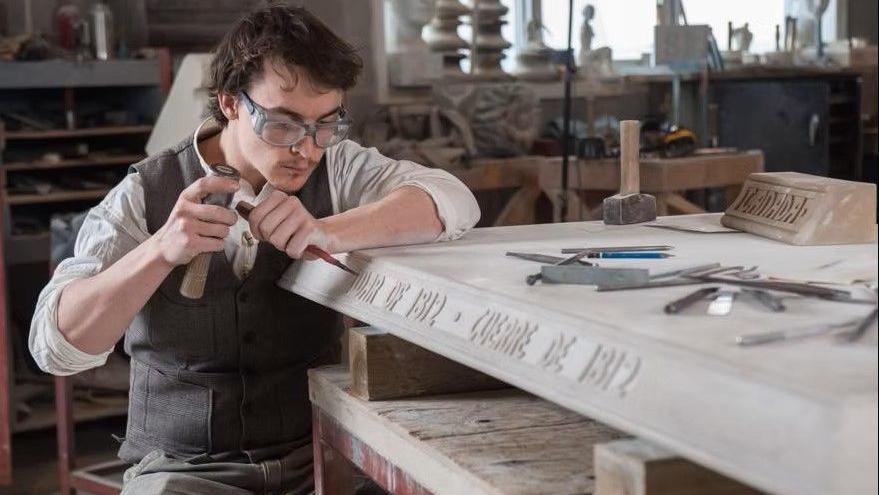 David Lamb carving an inscription on a piece of stone. He is sat by the flat piece of stone, holding carving tools. He has short brown hair and is wearing safety googles, a grey waistcoat jacked and cream shirt.