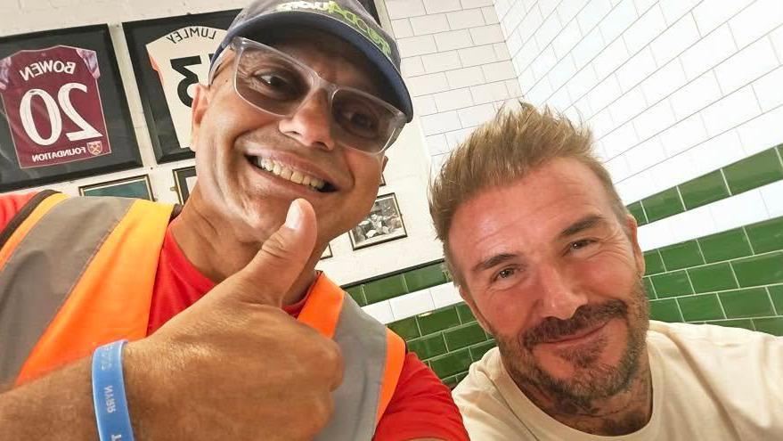 Pesh Kapashiawala is wearing an orange hi-vis vest over a red T-shirt. He is smiling and giving a thumbs up next to Sir David Beckham, who is wearing a white T-shirt and sitting down inside the restaurant. Sir David is also smiling.