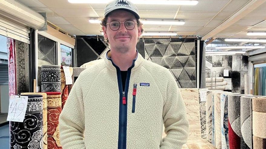 Joe Orr has a cream fleece, a light baseball cap and has round spectacles. He is standing in a carpet shop, with rolls of carpet standing on end behind him. 