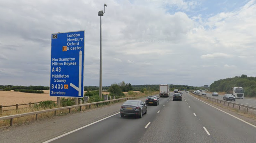 Junction 10 of the M40, a three-lane road. Cars can be seen driving along it. On the left, a blue sign shows an exit to the A43.