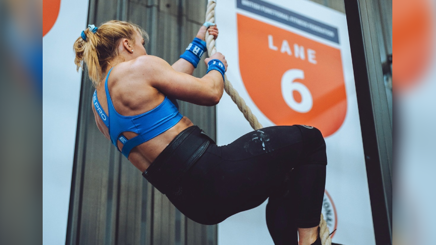 The picture shows Katherine Ogden climbing a rope. She is wearing a blue ves top and black leggings.