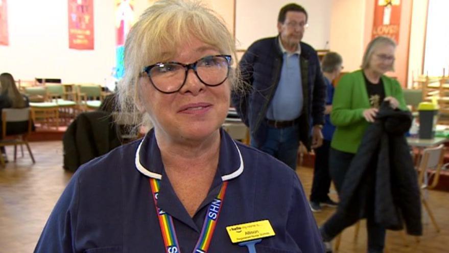 A blonde-haired woman with black-rimmed glasses and wearing a blue shirt with a yellow name tag that says Allison. 