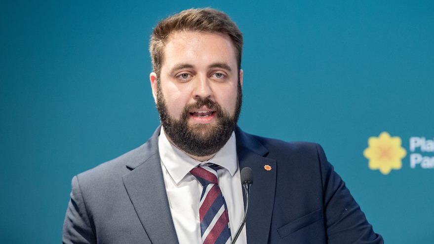 Luke Fletcher giving a speech at party conference in Cardiff, in front of a Plaid Cymru logo on a banner