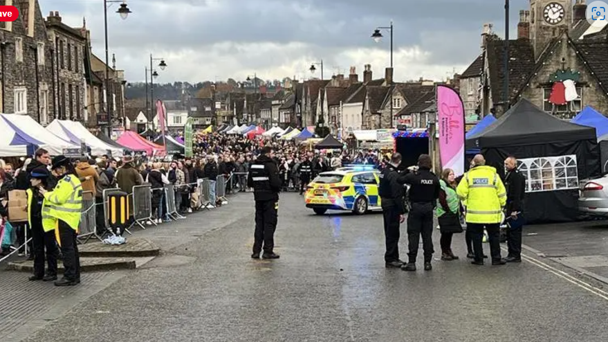 Chipping Sodbury High Street with several police officers and a police car in the foreground and lots of shoppers being kept away from the scene behind temporary barriers. Market stalls with coloured awnings stretch away into the distance