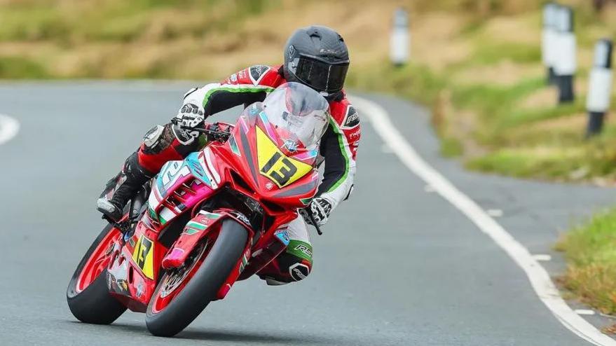 Louis O’Regan on a red bike with the number 13 on the front of it in black on a yellow plate. He is wearing red, green, black and white leathers and black helmet.