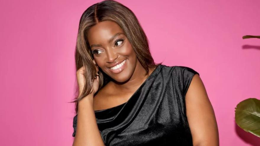 A woman wearing a black dress sitting on a chair in front of a pink background