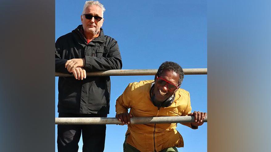 Henry Normal standing in front of railings and Lemn Sissay bending down and holding railings with blue sky