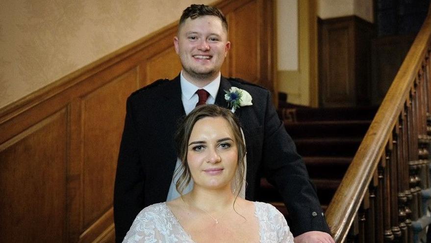 The couple pose on a stairway in the wedding venue which has wood-panelled walls and bannisters. Cameron stands behind his bride - he is wearing a kilt, jacket, burgundy tie and has a flower in his buttonhole while Weronika is in a lace gown and veil.