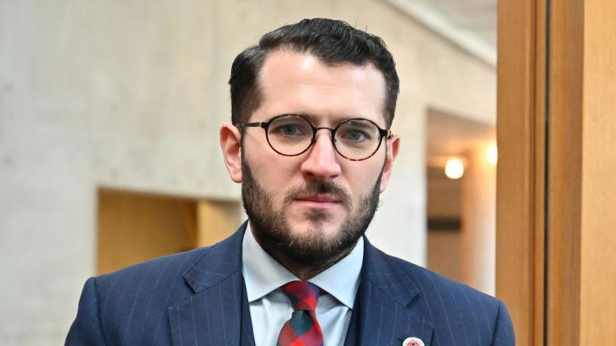 Paul Sweeney - a man with short dark hair, glasses  and a beard, wearing a dark blue suit.
