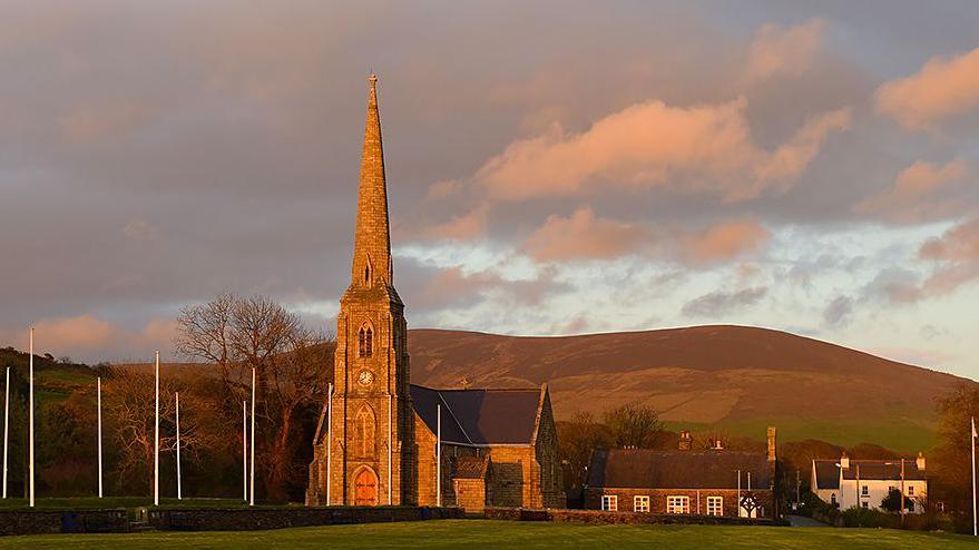The exterior of the Royal Chapel, a church with a thin, tall steeple, the afternoon sun makes it glow, and flag poles are in front, with hills in the background.