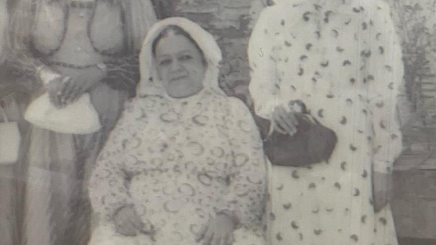 A black and white image of a woman sat down on a chair. Two other women are standing on either side of her, clutching handbags, but neither woman's face is visible. The seated woman has her hair tied back and is wearing a head covering. Her dress appears pale, with a circular print.