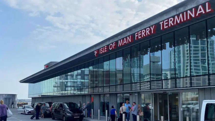 A long, two-story building with a glass frontage and an "Isle of Man Ferry Terminal" sign in large red and white letters along its roof. 
Passengers are standing outside and cars are also parked by the entrance. 