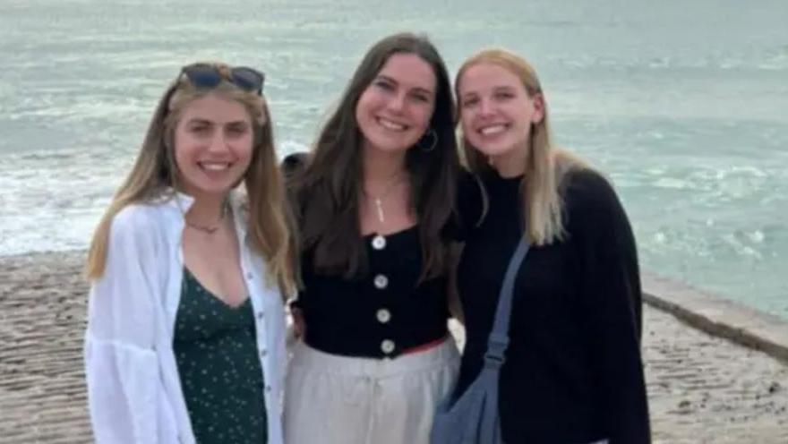 Three smiling women stand in front of a beach and coastline looking at the camera. the one on the left has blonde hair and glasses atop her head, she is wearing a green top and a white shirt. The girl in the middle has long dark hair a black top with white round buttons and a white pants, while the girl on the right has blonde shoulder-length hair and a black jumper, with a blue bag across her. They are huddled in together for the picture.