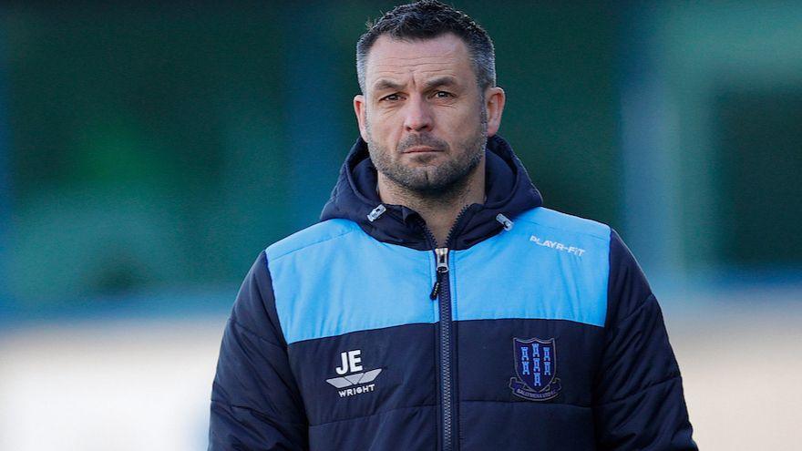 A man looks looks into the distance. He has short black hair which is greying at the sides. He has black and grey stubble. He is wearing a navy and sky blue coat with the Ballymena United crest. The background is blurred.
